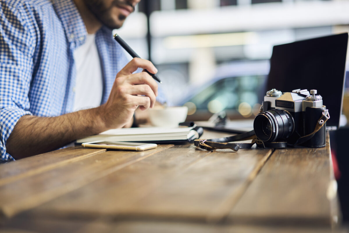 Cropped image of talented male photographer writing article about his retro exhibition of pictures made by vintage camera while working in modern coworking space sitting at wooden table with equipment